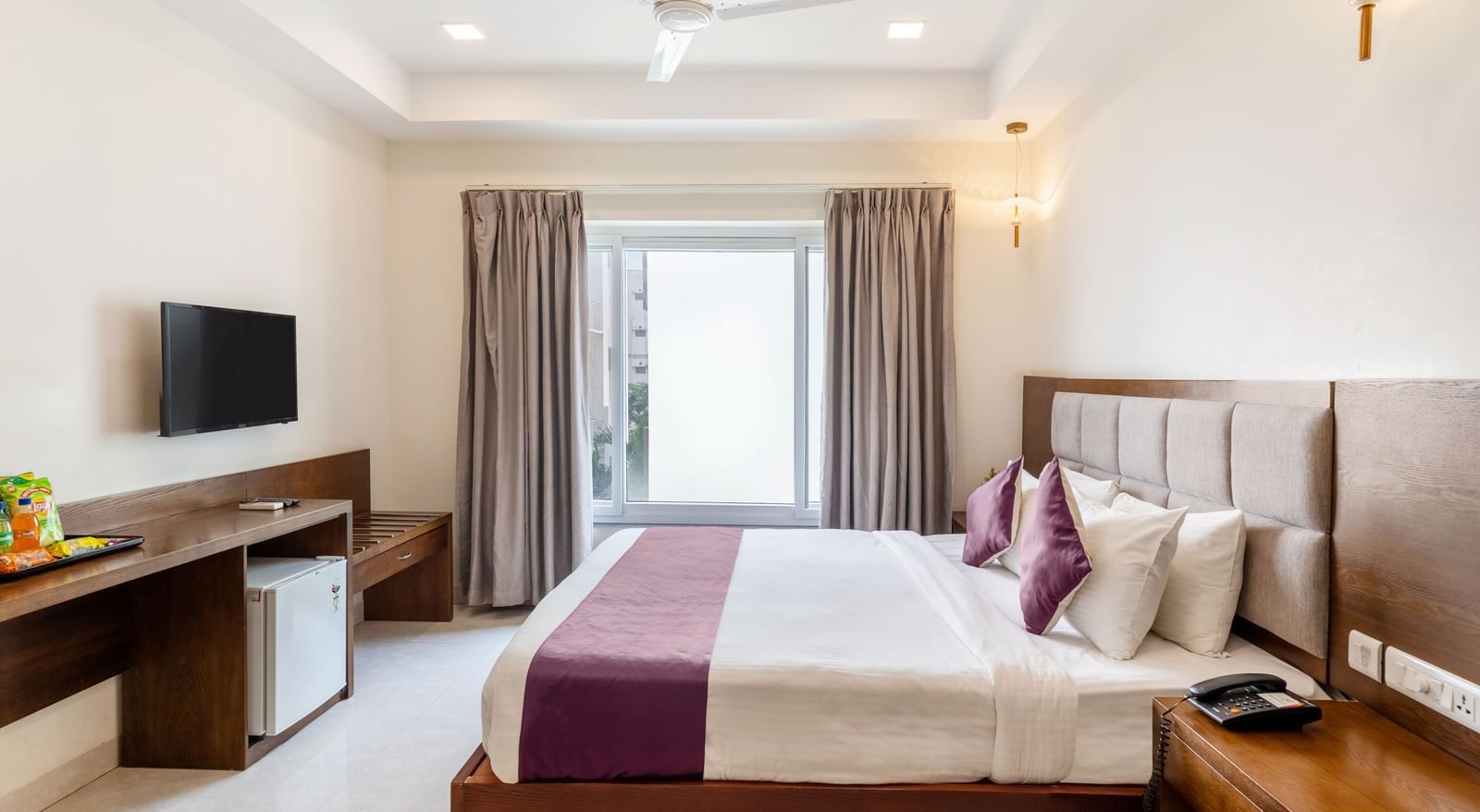 A well-lit room at Destino by Monday Hotels, Gachibowli, featuring a neatly made bed with a purple runner, a wooden desk with a minibar, and a large window.