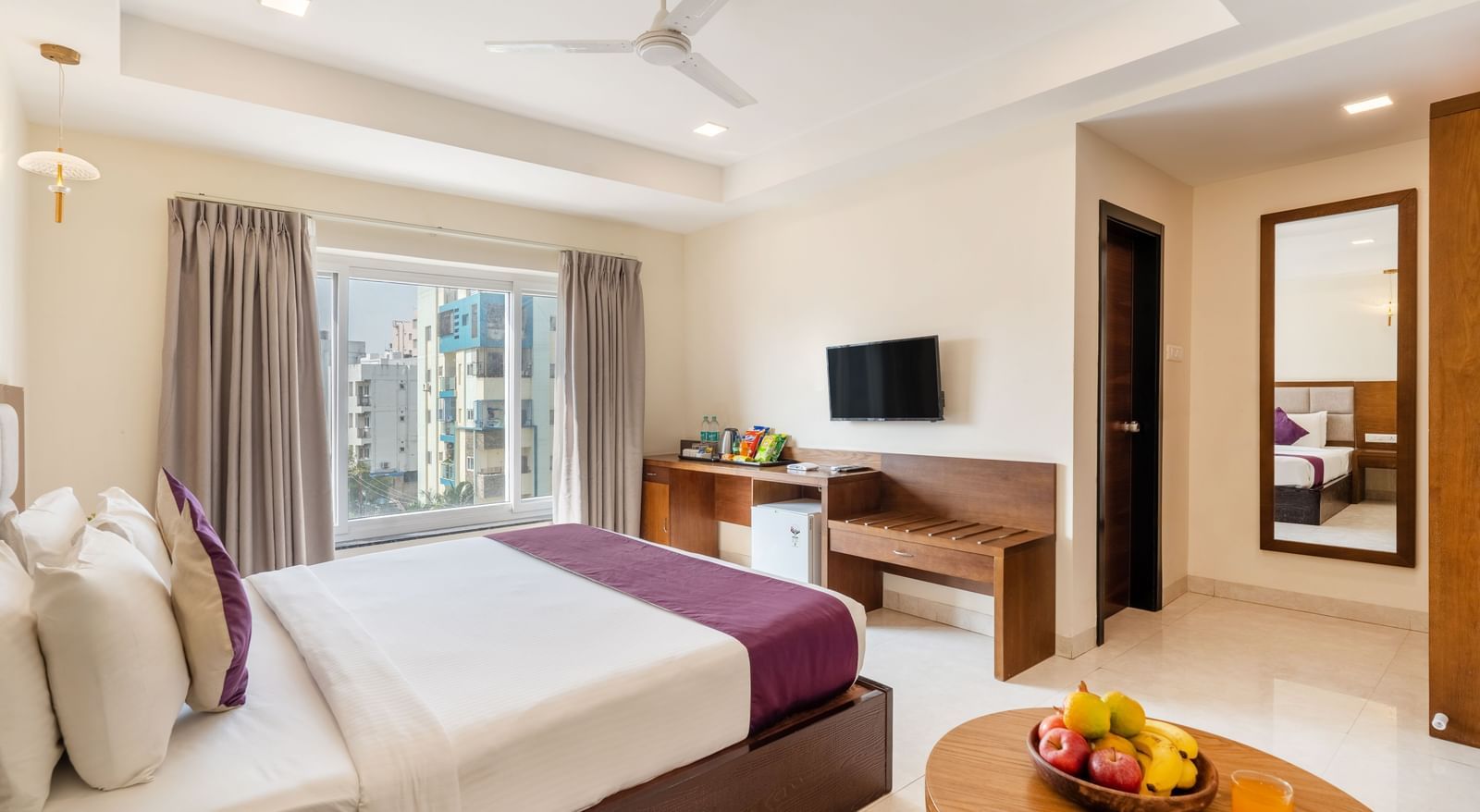 A spacious and well-lit room at Destino by Monday Hotels, Gachibowli, featuring a neatly made bed, a television, and a small round table with a fruit bowl.