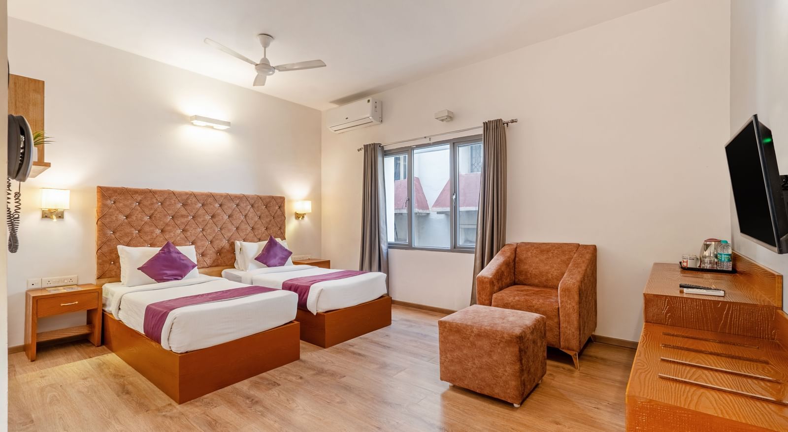 A bright hotel room with 2 single beds, cushioned headboards, a TV, a wooden desk, chairs, and a large window – Monday Hotels HITEC City, Hyderabad