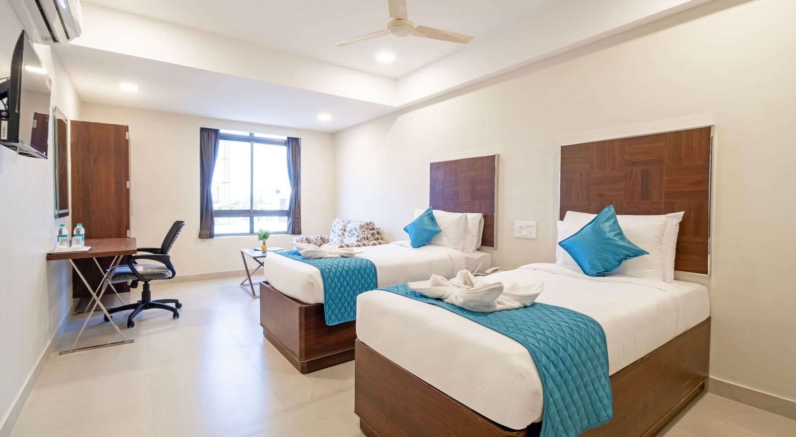 Twin beds with fresh linens blue runners seating area and bright ambience creating comfort at Swarna's The Capital.