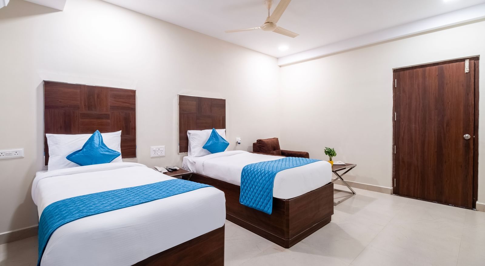 Twin beds with blue accents placed in a spacious bright room with wooden furniture and ceiling fan at Swarna's The Capital.