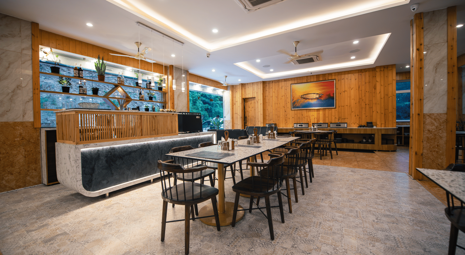 A restaurant at The Tattva Devaprayag with a long wooden table with black chairs and a bar.