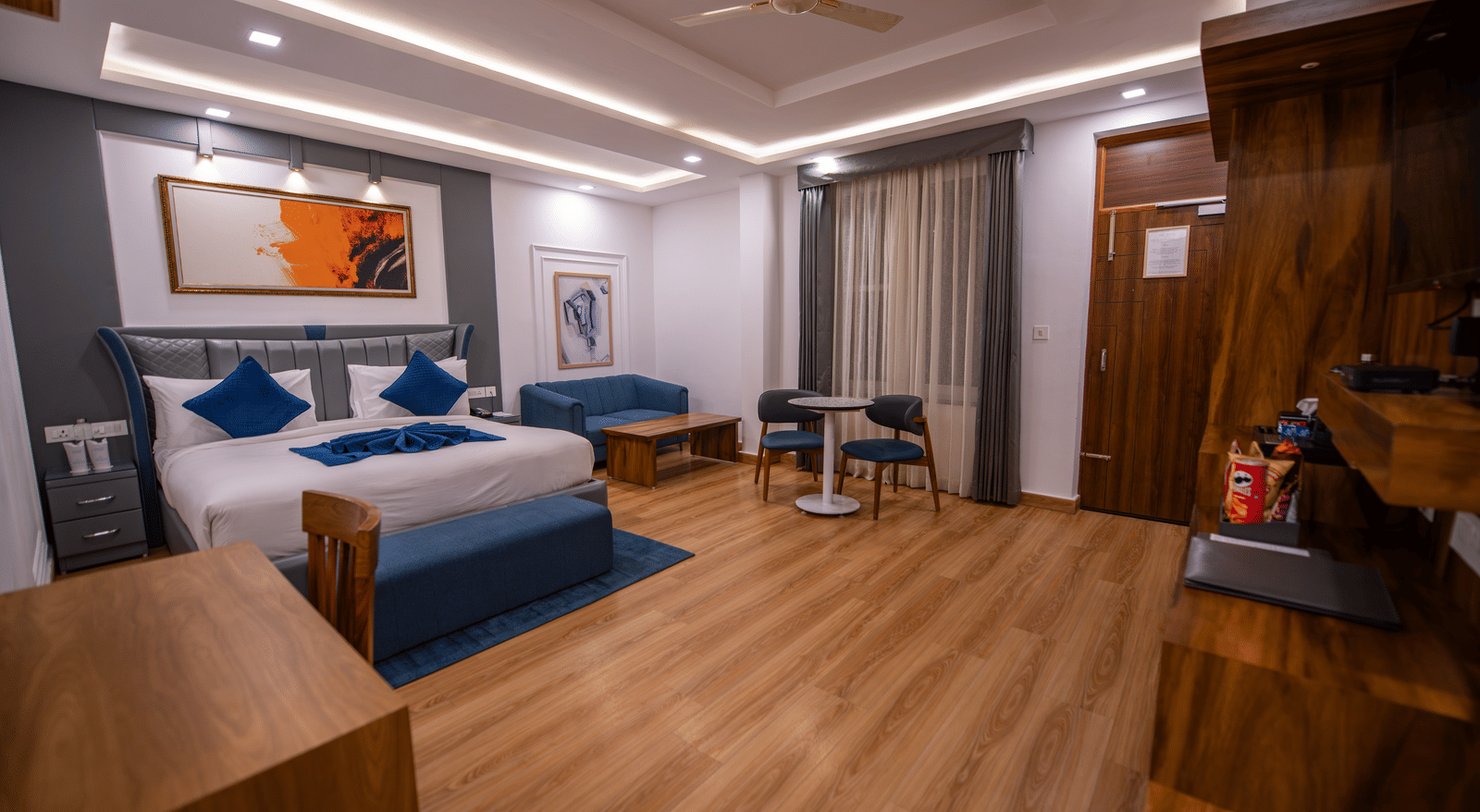 A superior room at The Tattva Devaprayag with a bed dressed in blue blankets and two pillows, a sofa, and framed paintings on the wall.