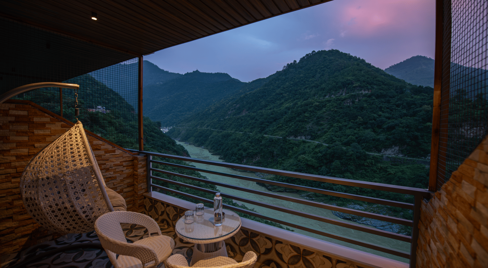 A shot of the mountains and river flowing from the balcony of The Tattva Devaprayag featuring two white chairs and a table during sunset.
