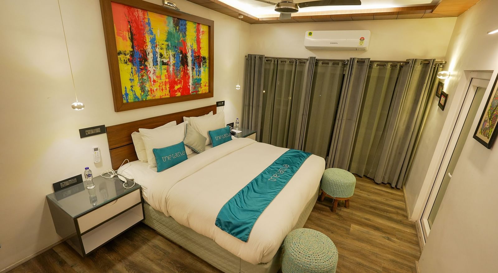 Bedroom of Pent House at The Tattva, Joshimath with a large bed, teal accents, abstract wall art, wooden flooring, grey curtains, and ambient ceiling lighting.