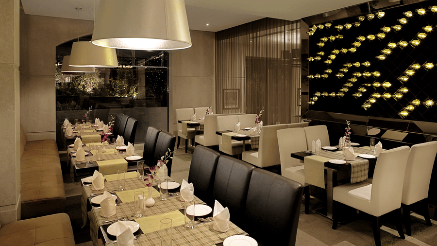 Modern dining room at Bahula restaurant in Barsana Boutique Hotel, Kolkata, featuring long set tables with elegant chairs and soft ambient lighting from large overhead lamps.