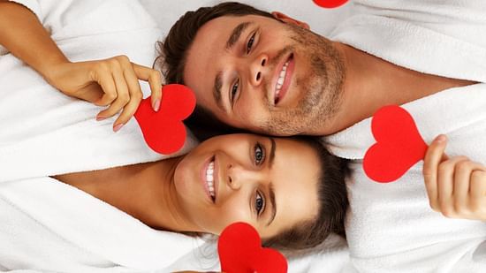 A smiling couple wearing white bathrobes holding red paper hearts on each hand, laying with their heads aligning adjacently opposite to each other's.
