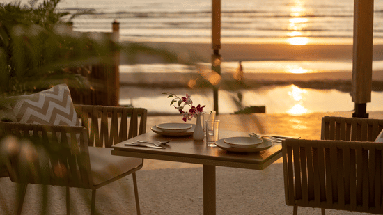 A close up shot of a table with two chairs at saz on the beach restaurant during sunset with sun rays reflecting on the sea waves in the background