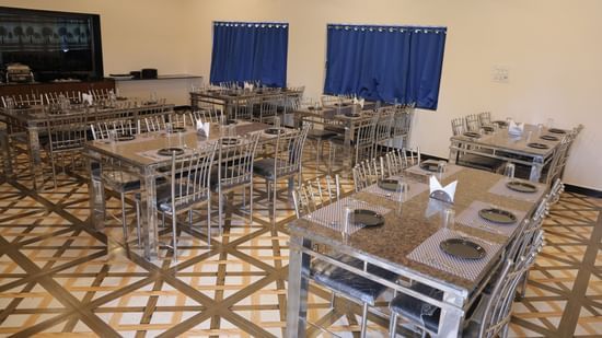A dining area with multiple tables and chairs, each set with plates, cutlery, and glasses. The tables are granite with steel frames, and there is a buffet counter in the background.