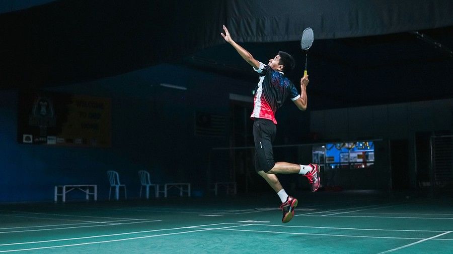 A male badminton player in mid-air performing a jump smash on an indoor court, wearing a black, red, and white sports outfit with red shoes, under focused lighting in a dimly lit gym.