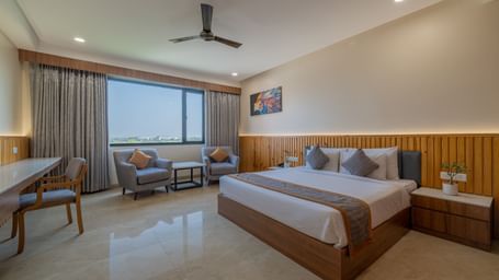 A bedroom at Hotel Highway King, Bagru, featuring bedside tables, desk, seating area, ceiling fan, and large window.