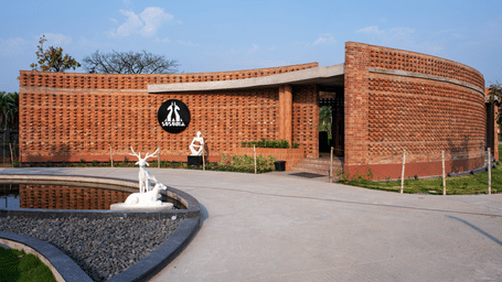 The curved brick entrance of the reception with landscaped pathways and a decorative fountain at the driveway entrance under a clear blue sky | Hotel Sonar Bangla, Susunia