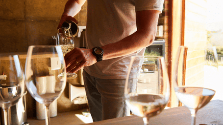 A man filling several wine glasses with white wine in a shed with ample of natural light coming from the outside.