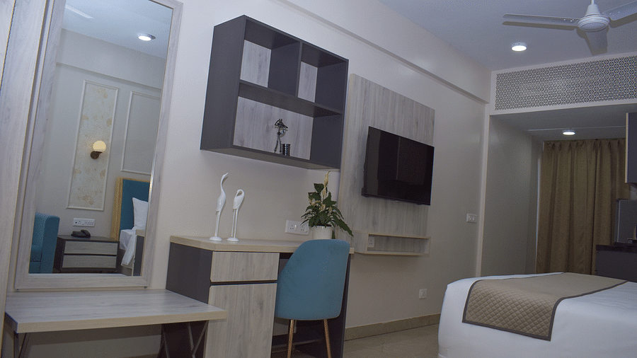 A hotel room with a desk and chair, a wall-mounted TV, a wall cabinet, a mirror, and a partial view of a bed and kitchenette area - Urban Suites by BluSalzz, Jaipur