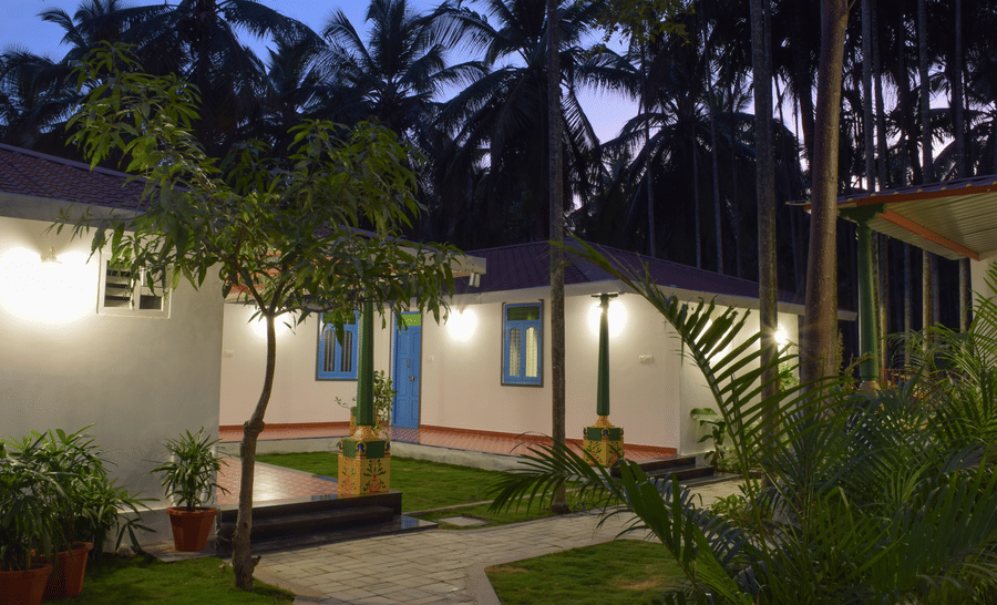 A white resort cottage with a red roof and blue window frames, surrounded by palm trees and green lawns - Ibex Resorts, Coimbatore (Kakarla)