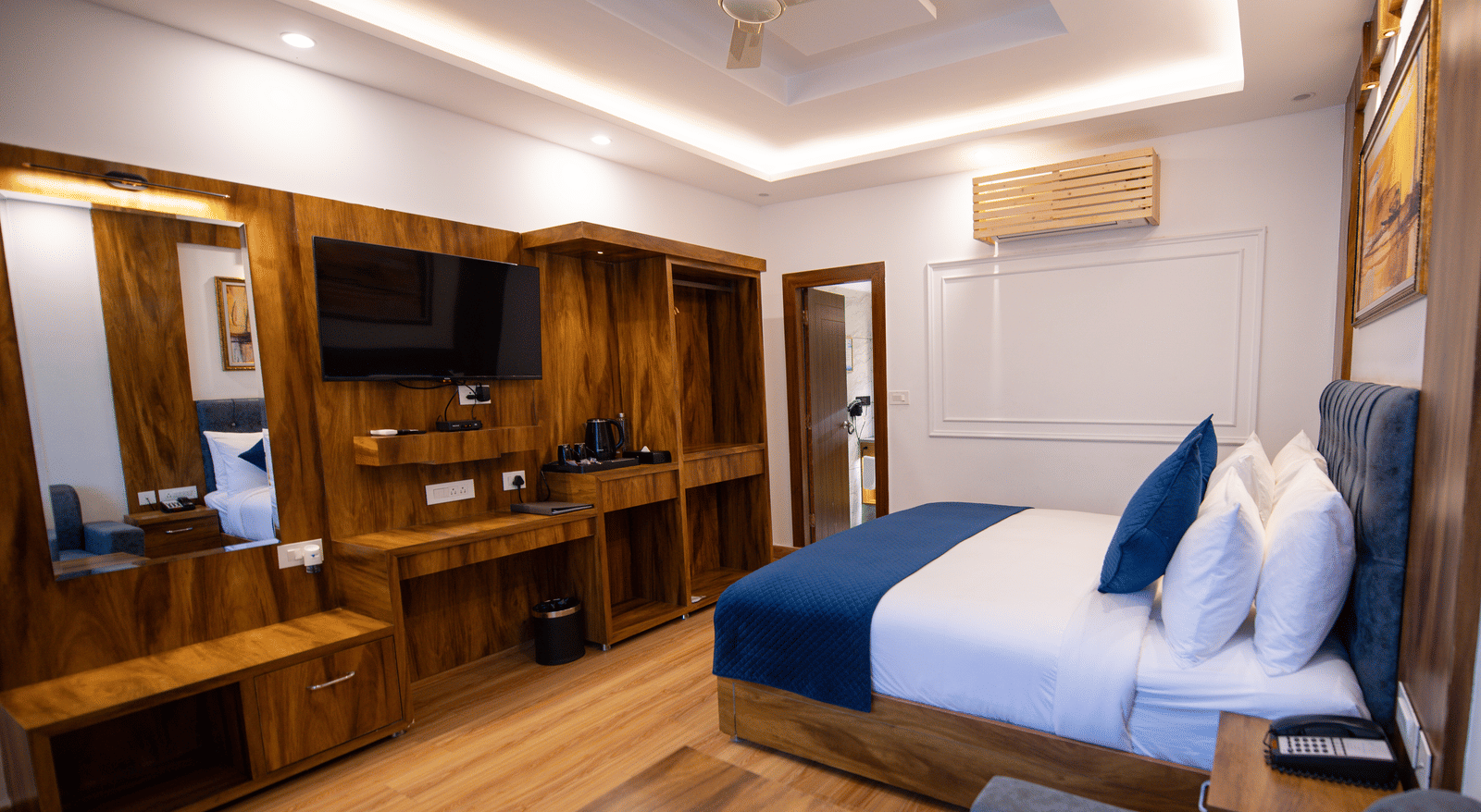 A Deluxe Room at The Tattva Devaprayag with a bed featuring blue blankets and two pillows, a desk, a television, and a sofa.