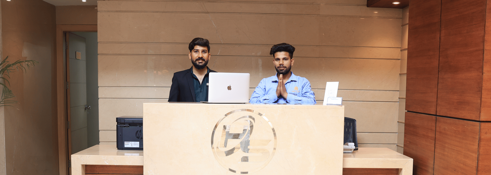 A hotel reception desk made of wood and light stone, with two male staff members standing behind it | Perfectstayz Value Sagar