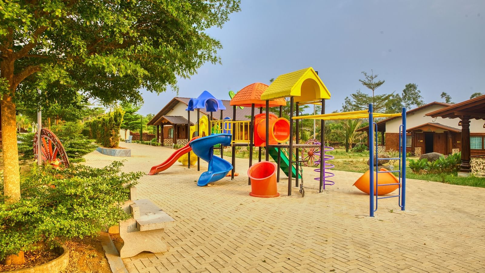 A colourful outdoor children's playground with various slides, climbing frames, and swings on a paved area, surrounded by trees. - Vintage Trails by Ilara
