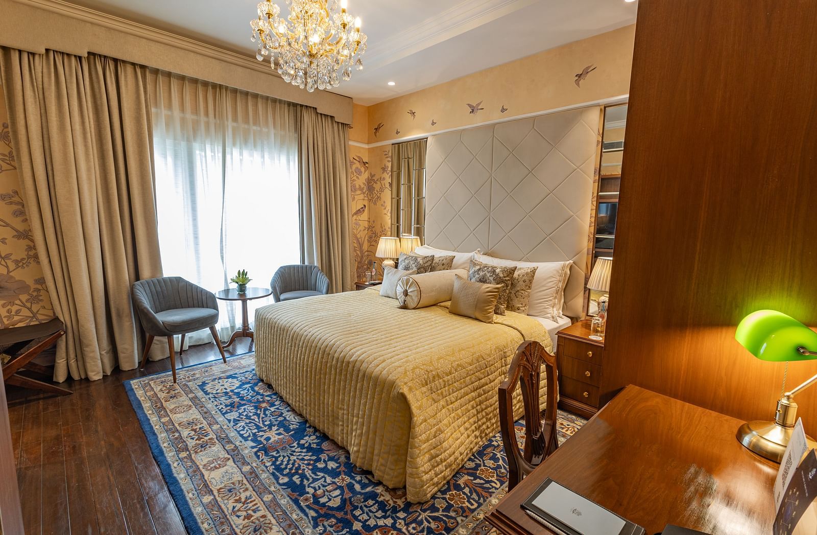 Luxury King Bed Room with Balcony at The Claridges New Delhi, featuring a cosy bed with pillows, a glass door covered by curtains, and a chandelier hanging overhead