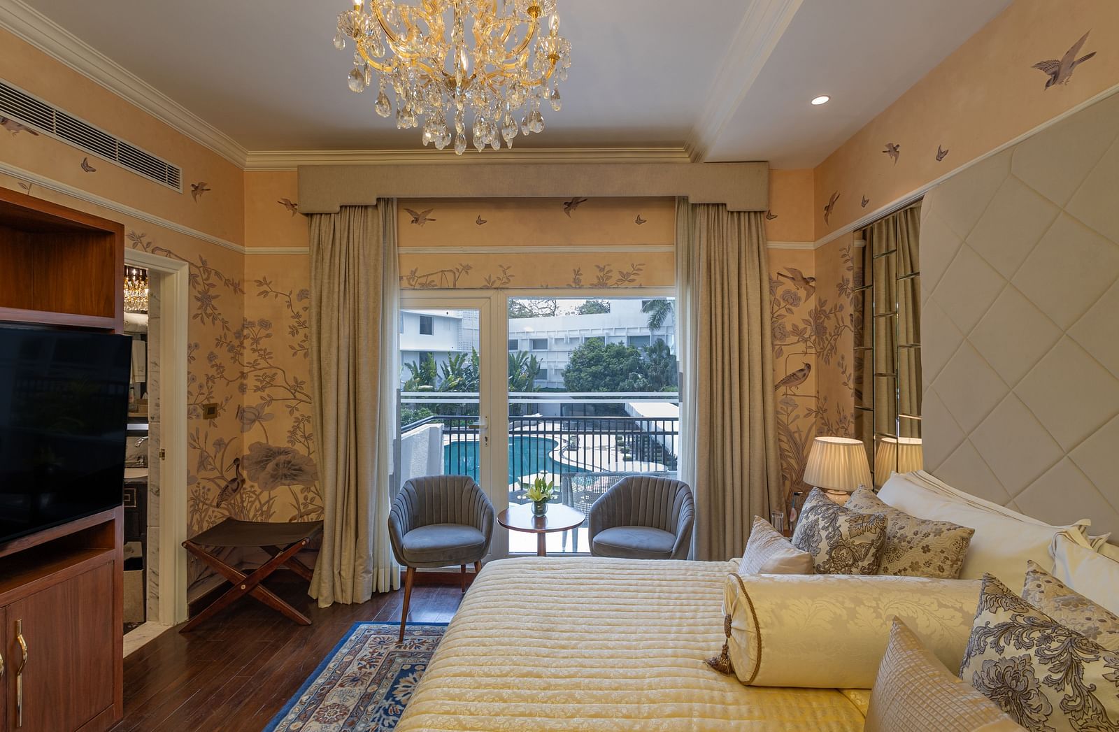 Side view of Luxury King Bed Room with Balcony at The Claridges New Delhi, featuring a cosy bed with pillows, a glass door covered by curtains, and a chandelier hanging overhead