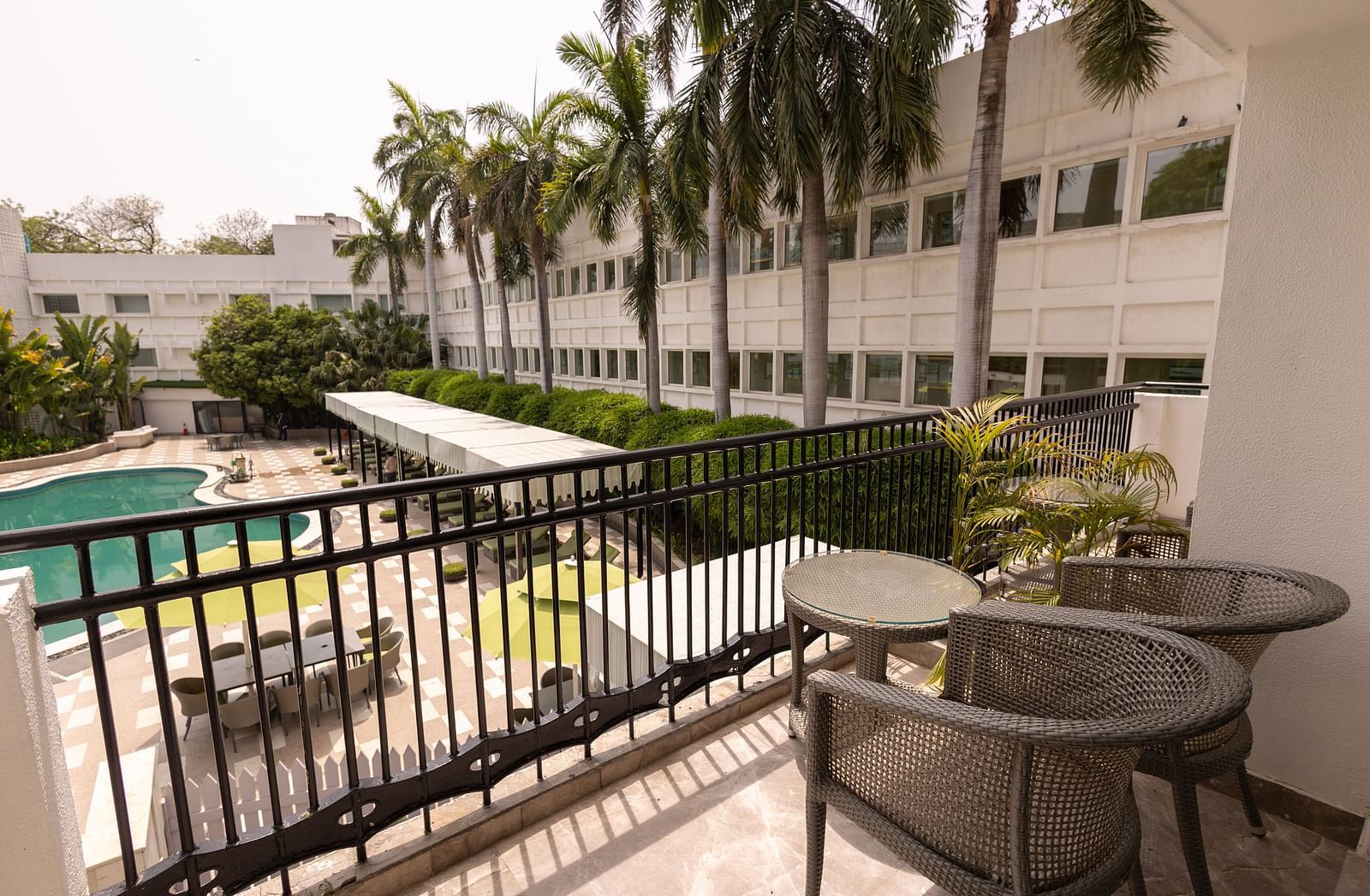Luxury King Bed Room with Balcony at The Claridges New Delhi, offering a view of the hotel’s outdoor open-air swimming pool surrounded by lush trees and nearby buildings
