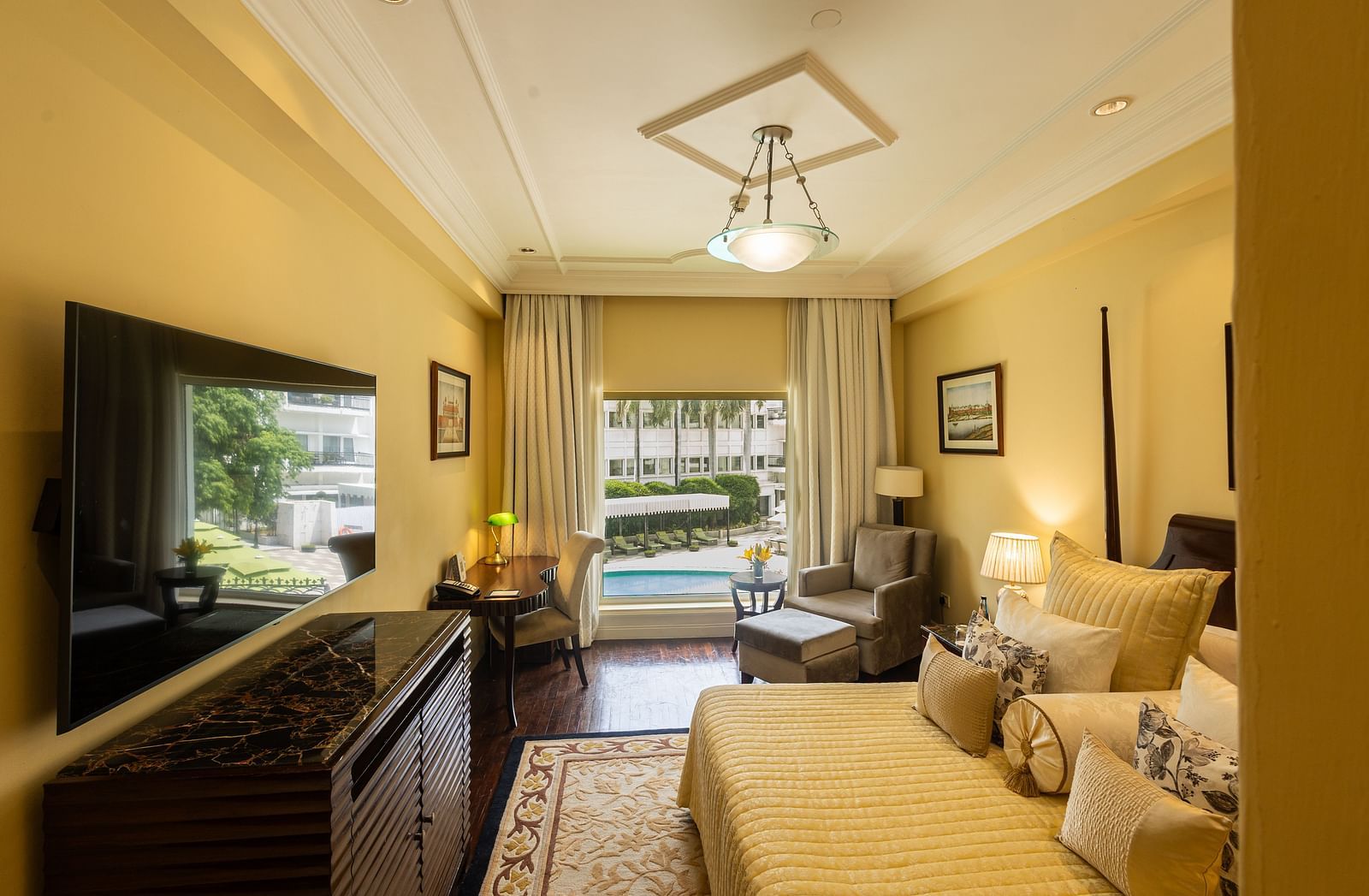 Side view of the Deluxe King Bed Room Pool View at The Claridges New Delhi, featuring a cosy king sized bed, wall-mounted TV, a corner chair, and a large window overlooking the swimming pool