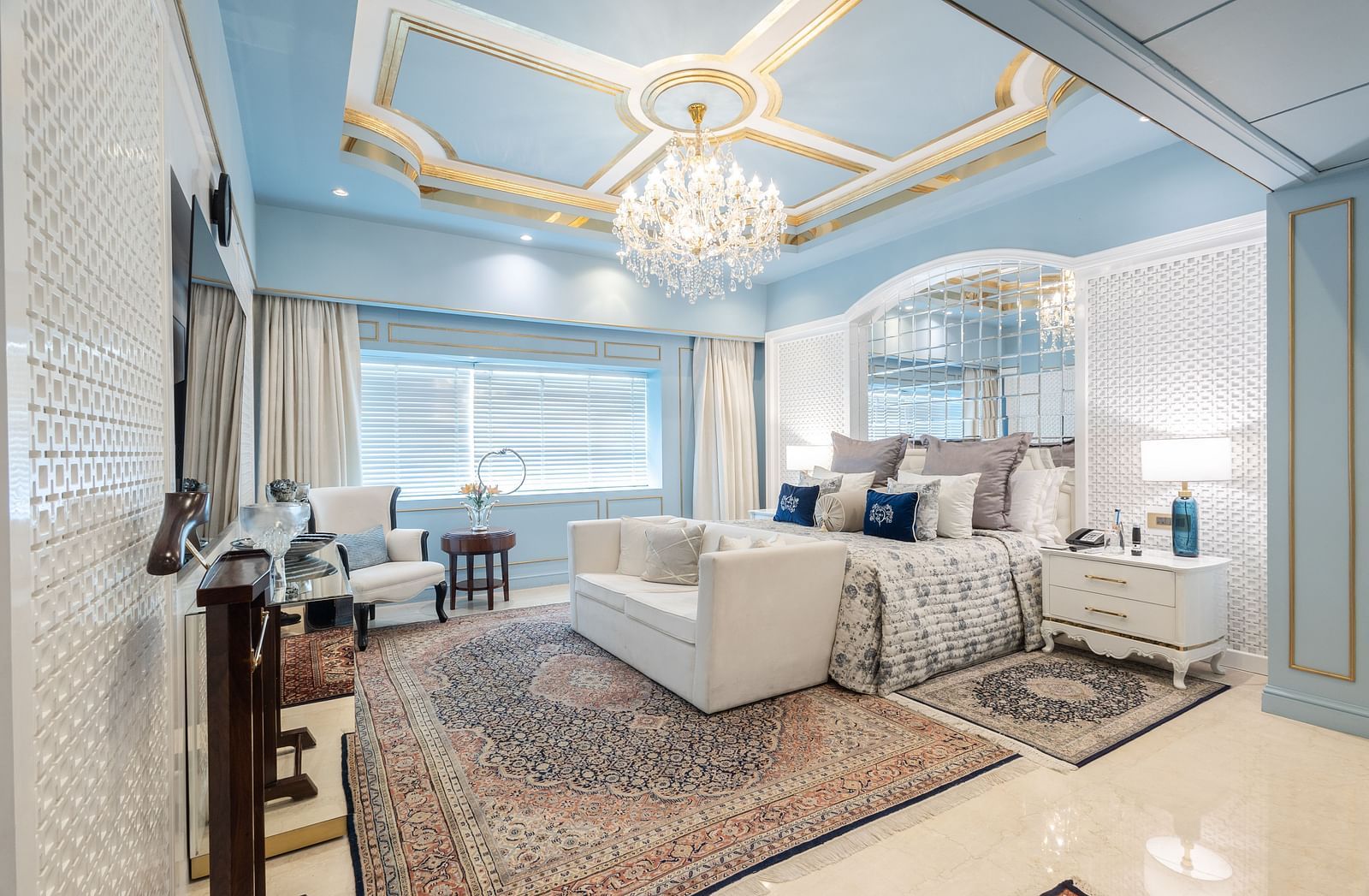 An image of a suite with a large bed and a sofa placed in front of a television and a chandelier hanging from the ceiling