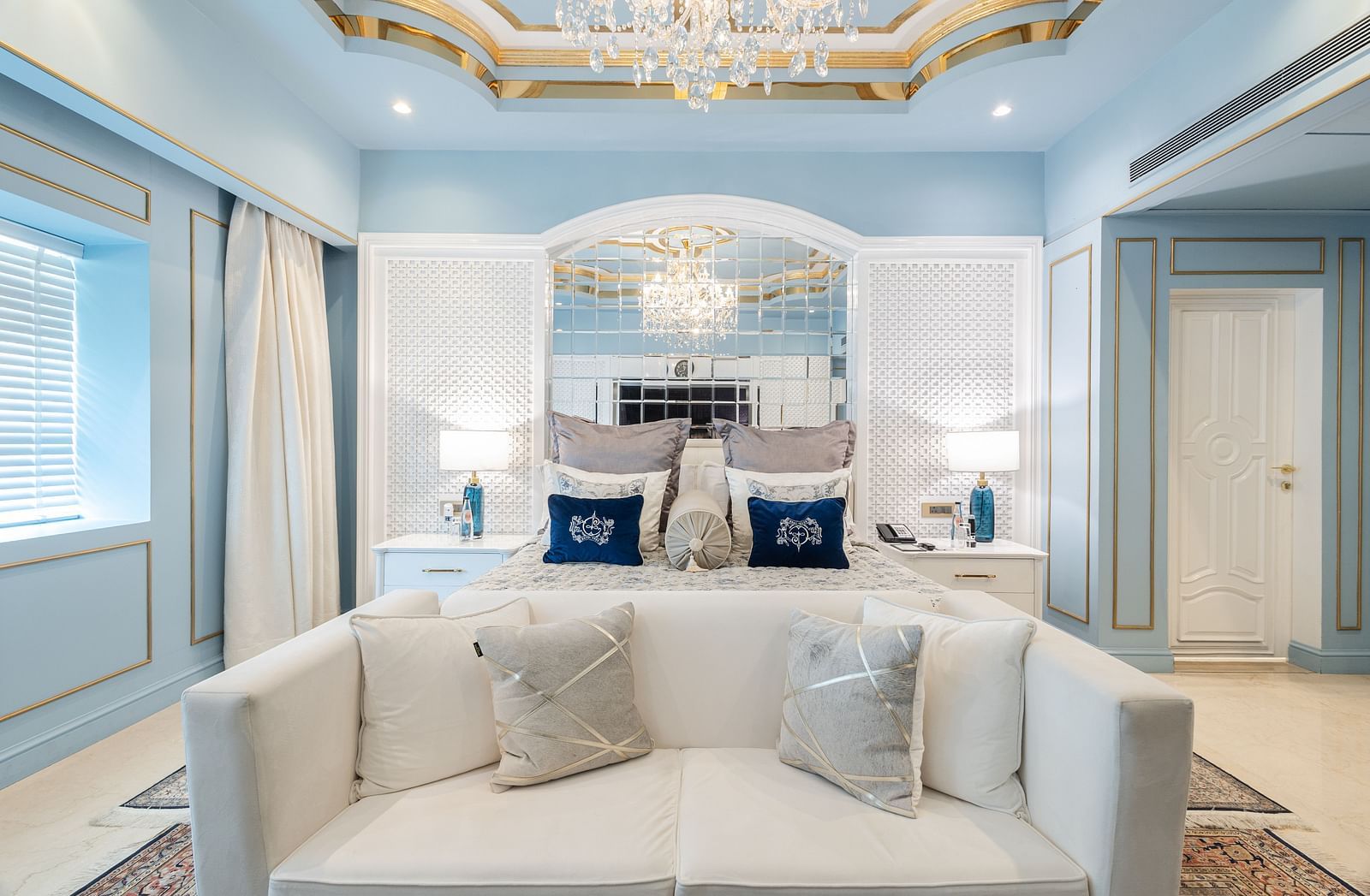 An image of a couch with cushions placed in front of a bed under a chandelier in our Diplomatic suite room - The Claridges New Delhi