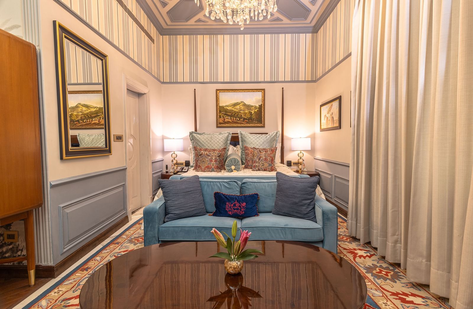 image of the bedroom of the heritage parlour suite with a king sized bed and a blue couch with two cushions placed on it - The Claridges New Delhi