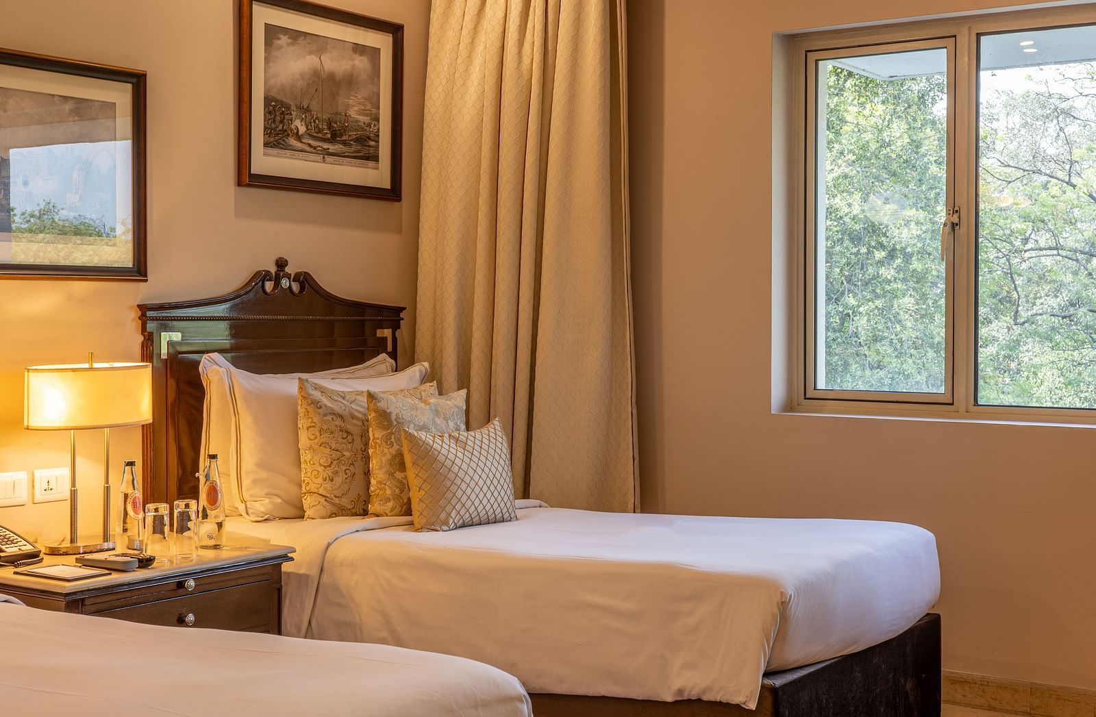 Grand Luxury Twin Bed Room at The Claridges New Delhi, featuring elegant twin beds, framed photos on the wall, a bedside lamp between the beds, and a window