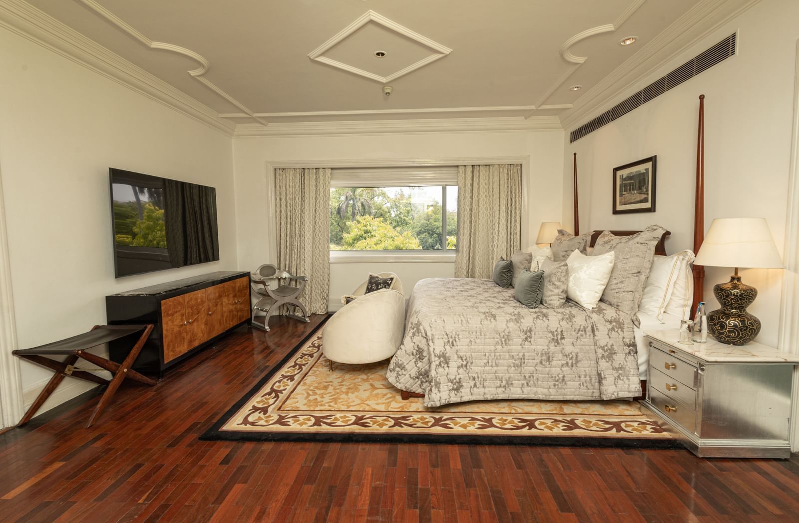 The Claridges New Delhi spacious bedroom has dark wood flooring, a patterned bedspread, and large windows.