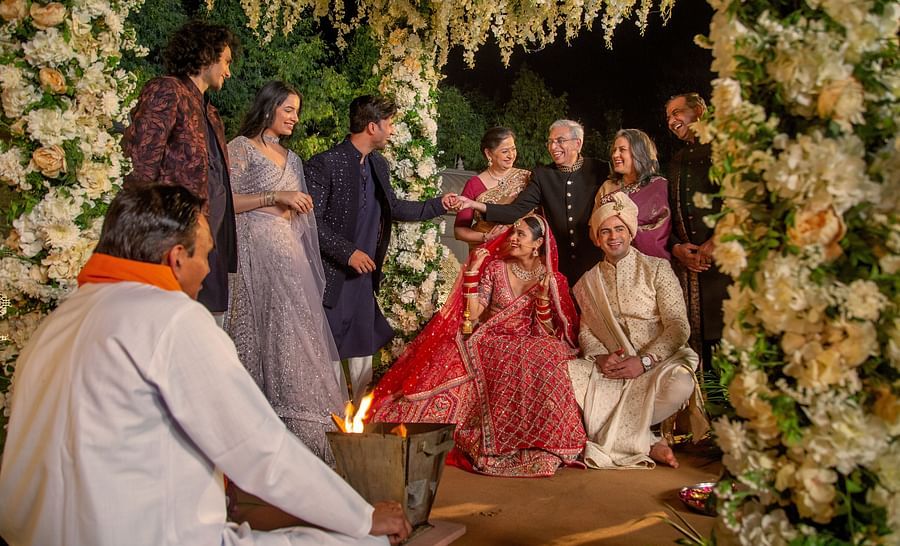A traditional Indian wedding ceremony with a bride in a red dress sitting beside the groom, surrounded by family and a lush floral backdrop.
