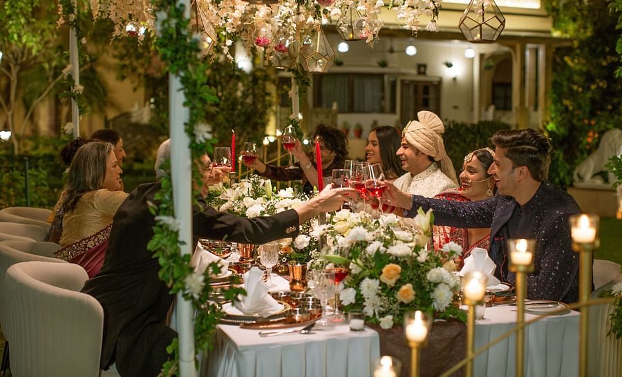 An elegant dinner party scene with guests seated around a table lavishly decorated with flowers and candles, passing dishes in a warmly lit garden setting.