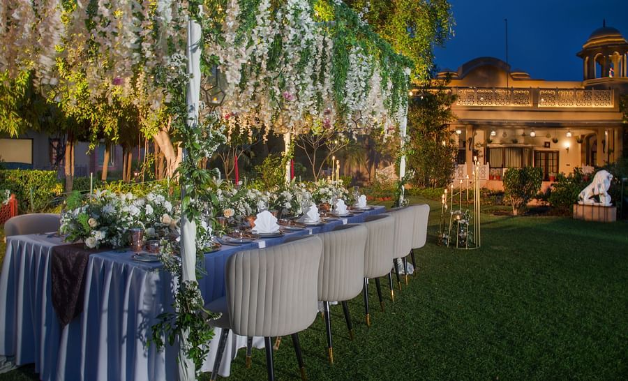 An elegant outdoor dining setup at night, featuring a long table with white tableware and floral arrangements, under cascading fairy lights and surrounded by lush greenery.