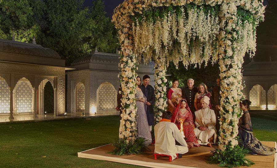 A traditional indian wedding ceremony at night, with the bride and groom under a beautifully decorated floral canopy, surrounded by guests in formal attire, set against a backdrop of ornate arches.