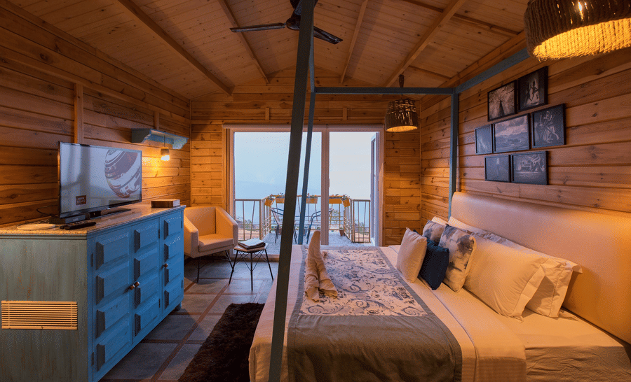 A bedroom at Polo Cherrapunjee Resort with a bed, a colourful dresser, seating area, and glass doors opening to a balcony with scenic views.