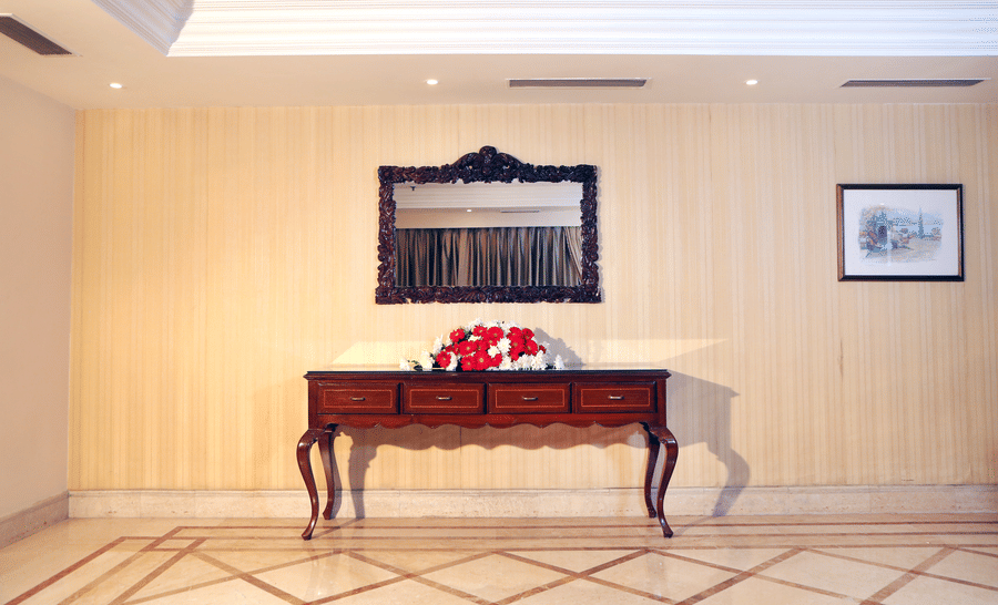 Hallway at The Bristol Hotel, Gurgaon with a wooden console table, an ornate carved mirror, a red and white flower arrangement, and a framed painting.