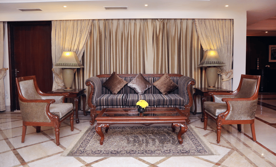 Lounge seating area at The Bristol Hotel, Gurgaon with a carved wooden sofa, 2 matching armchairs, a glass-top coffee table, table lamps, and gold curtains.