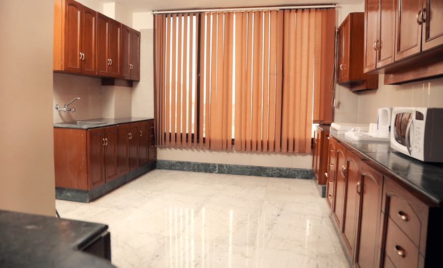 Kitchenette at The Bristol Hotel, Gurgaon with wooden cabinetry, a sink, a microwave, vertical blinds over the window, and marble flooring.