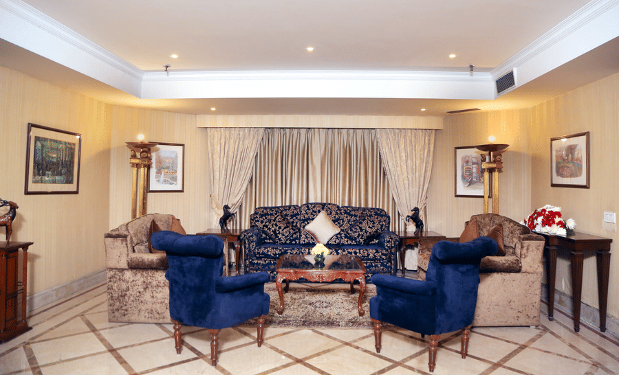 Suite living room at The Bristol Hotel, Gurgaon with a navy floral sofa, 2 blue velvet wingback chairs, a wooden coffee table, gold curtains, and framed art.
