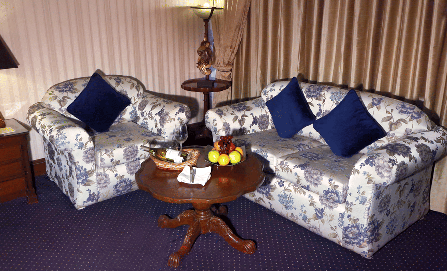 Suite sitting area at The Bristol Hotel, Gurgaon with 2 floral-print sofas, navy cushions, a round wooden table with a fruit basket, and a bottle of wine.