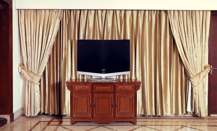 Television console at The Bristol Hotel, Gurgaon with a flat-screen TV on a wooden cabinet with 3 cupboard doors, set against floor-length gold curtains.