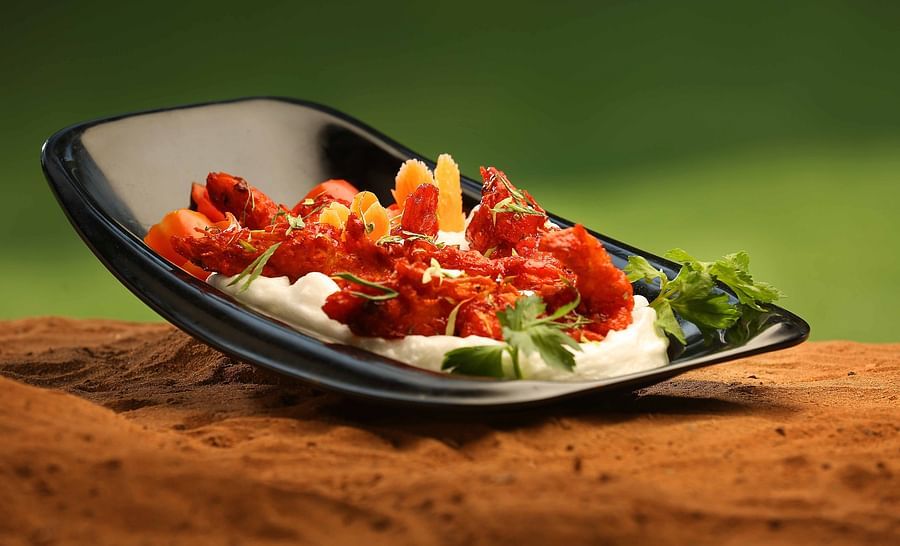 A black, boat-shaped plate on a textured surface holds a vibrant red dish over a white creamy base, garnished with orange slices and fresh parsley - Uday Samudra Leisure Beach Hotel.