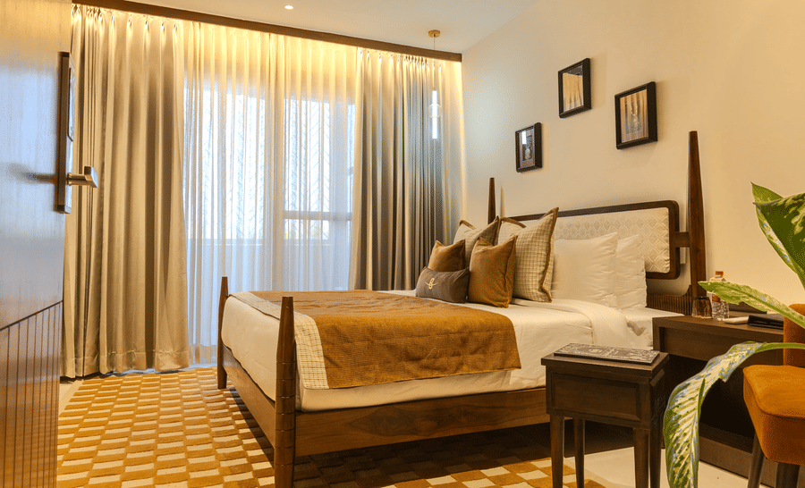 Spacious bedroom with a king size bed dressed in warm toned linen, wooden headboard and soft curtains at Hotel Bawa, Udaipur.