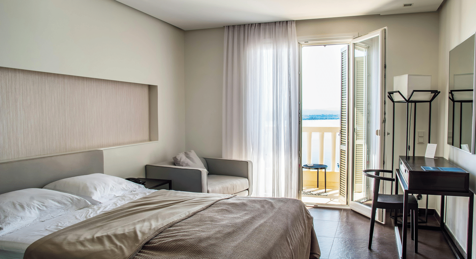 A modern room featuring a doule bed, wooden furniture, and a balcony door overlooking the ocean.