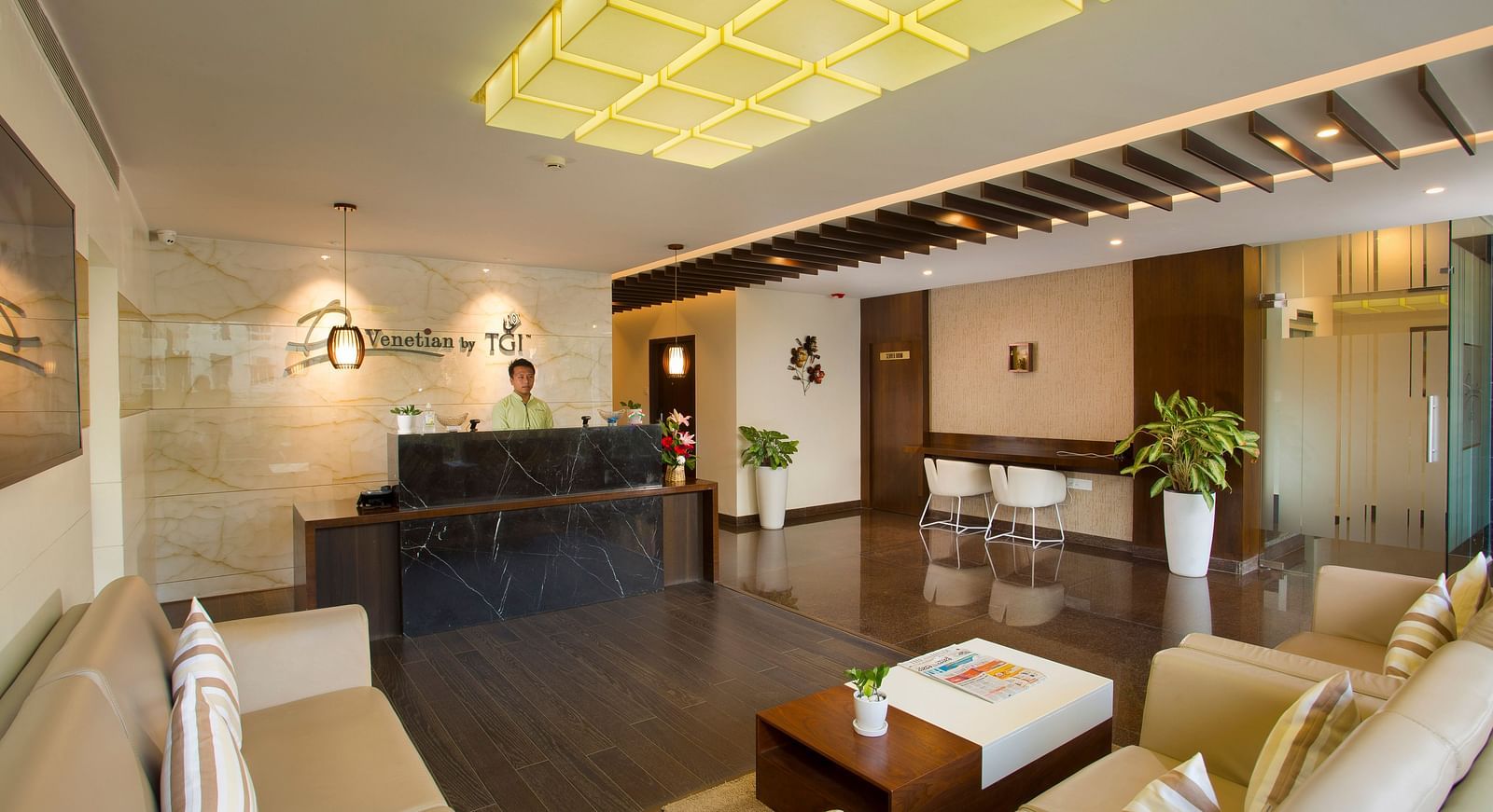 A hotel lobby featuring a marble reception desk, modern seating and geometric ceiling lights - De Venetian by TGI, Brookefield