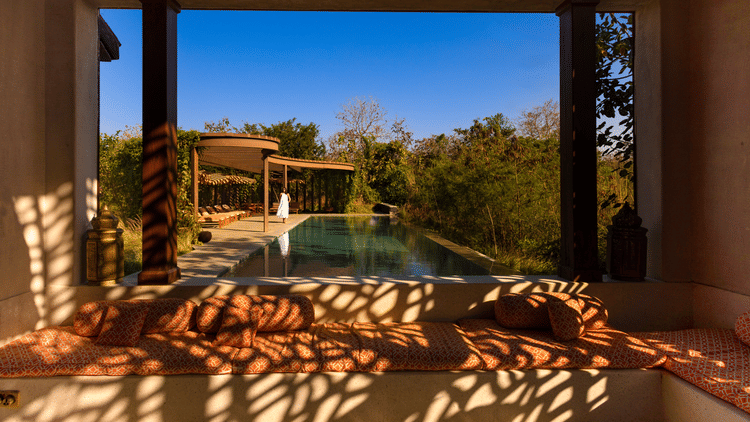 Open lounge area at Aramness, with dappled sunlight creating intricate shadow patterns on the seating and walls, overlooking a private swimming pool surrounded by lush greenery and forest views.