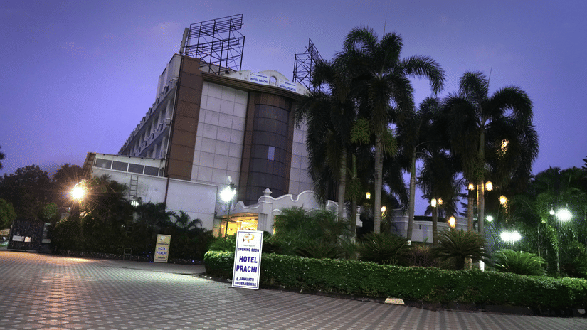 Facade of Sarovar Portico, Bhubaneswar, seen under the dark sky, with a well-lit driveway, a manicured garden, and a board with the name of the hotel on it.