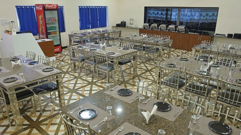 A wide shot of the restaurant at VITS Select Diamond Resort, Sasan Gir. The room is filled with tables and steel chairs, a buffet counter, and a Coca-Cola refrigerator.
