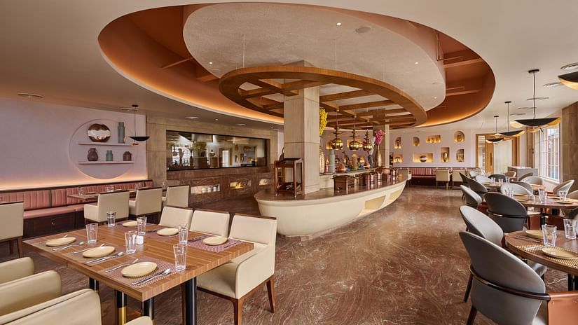 A stylish restaurant interior showcases a centrally located, elliptical bar with hanging pendant lights. Dining tables with set places are arranged throughout the space, which features a patterned marble floor and warm lighting - Voyage The Solitaire Bliss